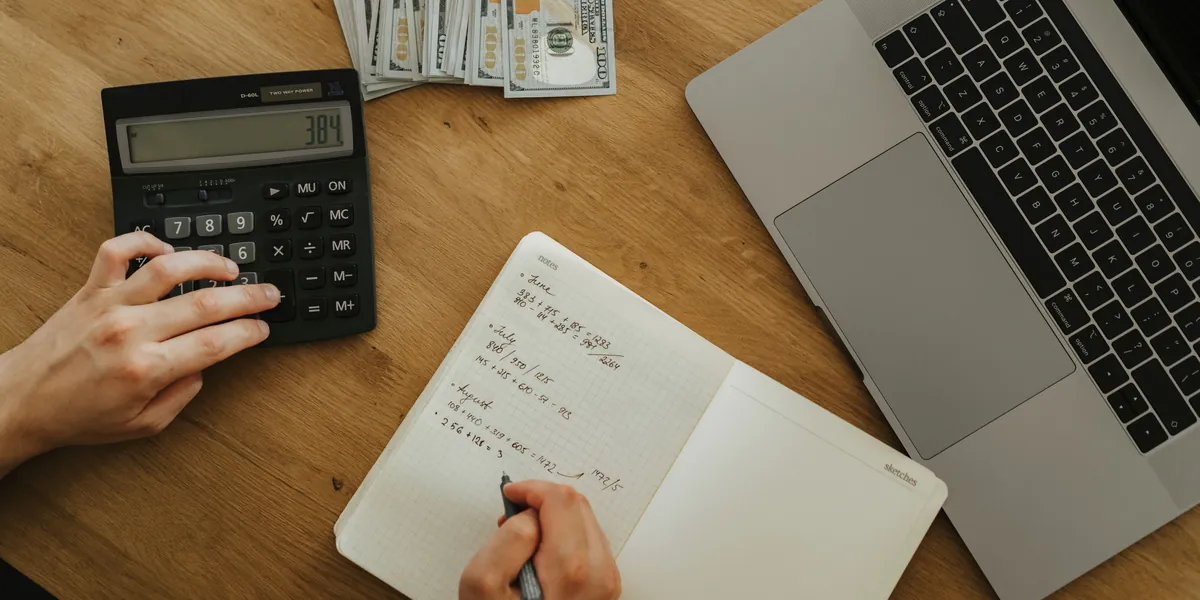 Mâini care tastează pe un calculator negru lângă un caiet cu socoteli lunare scrise de mână, un laptop și câteva bancnote, văzute de sus