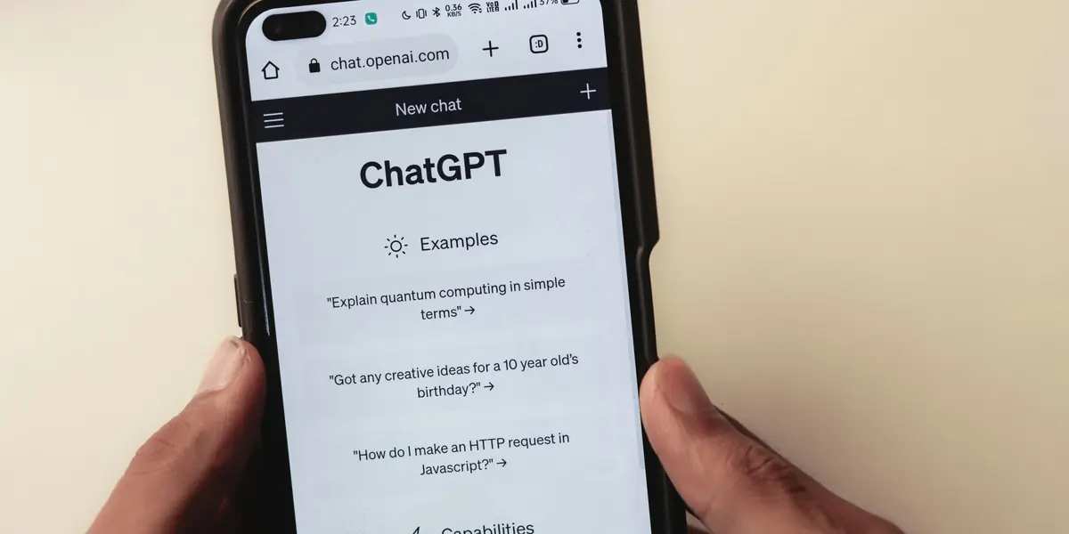 Two hands holding a phone in front of a cream wall, the browser open at chat.openai.com showing a New chat screen headed ChatGPT with example prompts including Explain quantum computing in simple terms and How do I make an HTTP request in Javascript