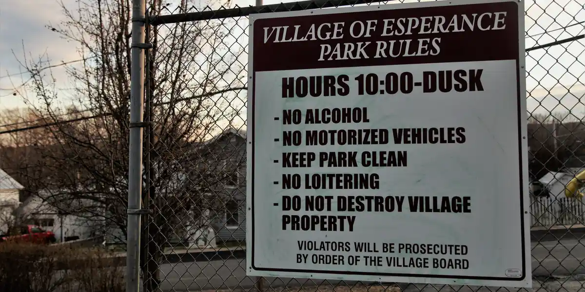 Panou alb prins de un gard de sârmă, cu textul VILLAGE OF ESPERANCE PARK RULES, HOURS 10:00-DUSK, NO ALCOHOL, NO MOTORIZED VEHICLES, KEEP PARK CLEAN, NO LOITERING, DO NOT DESTROY VILLAGE PROPERTY, VIOLATORS WILL BE PROSECUTED BY ORDER OF THE VILLAGE BOARD