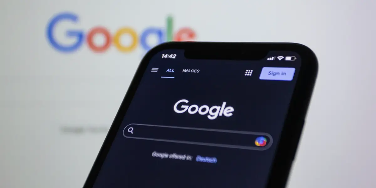 Uno smartphone con l'app Google aperta, tenuto davanti a un monitor che mostra la stessa pagina di ricerca
