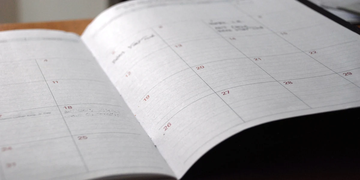 Agenda mensile aperta su un tavolo di legno, con i giorni 11, 18, 25 sulla pagina sinistra e 13, 19, 20, 21, 22, 26, 27, 28, 29 su quella destra, gli appunti a mano illeggibili