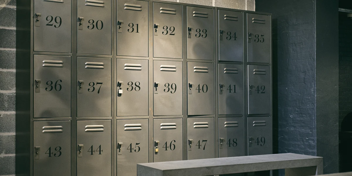 Tre file di armadietti metallici grigi numerati dal 29 al 49, con serrature e due lucchetti agganciati, davanti a un muro di mattoni e a una panca di cemento