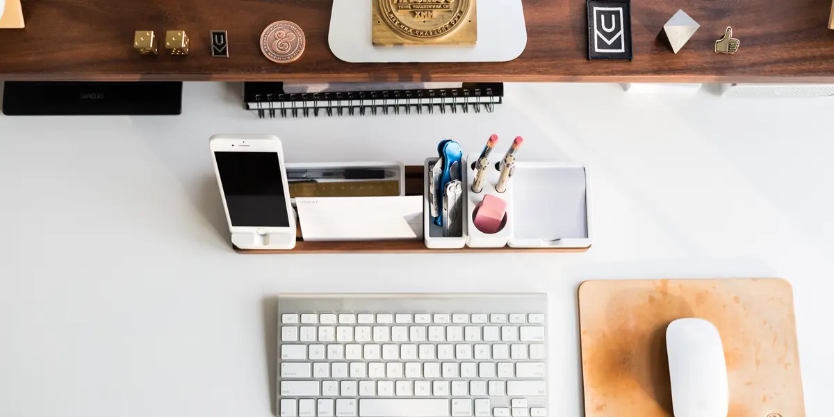 Overhead view of a workspace on a white top with a wooden shelf: pale keyboard, mouse on a cork mat, phone in a stand, pens and small desk objects
