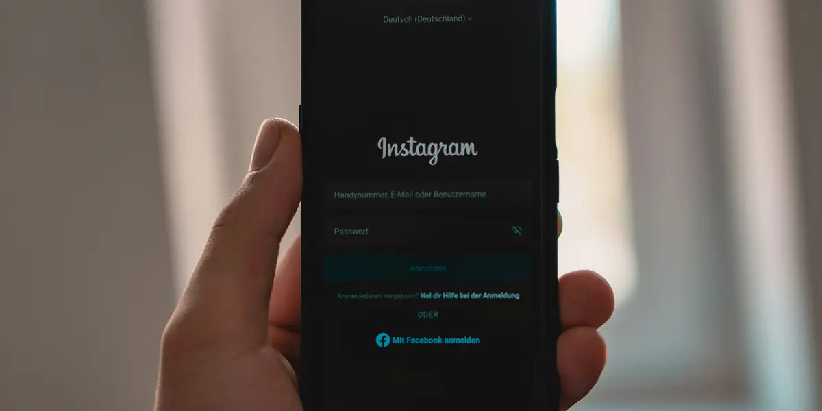Mână care ține un telefon cu ecranul de autentificare Instagram în limba germană: câmpuri pentru număr de telefon sau nume de utilizator și pentru parolă, iar dedesubt butonul albastru Mit Facebook anmelden pentru intrarea cu contul de Facebook